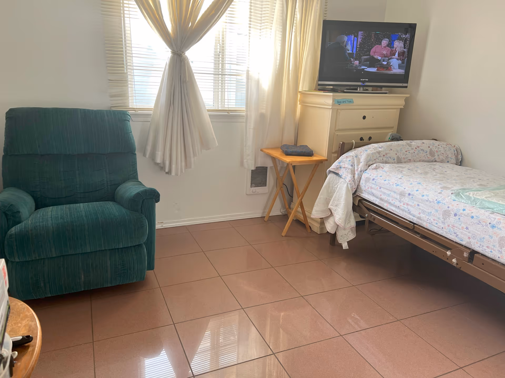 A small bedroom with a single bed covered in a floral patterned blanket, a green upholstered armchair, a wooden side table with a folded towel on it, a cream-colored dresser with a television on top, and a window with white curtains letting in natural light.