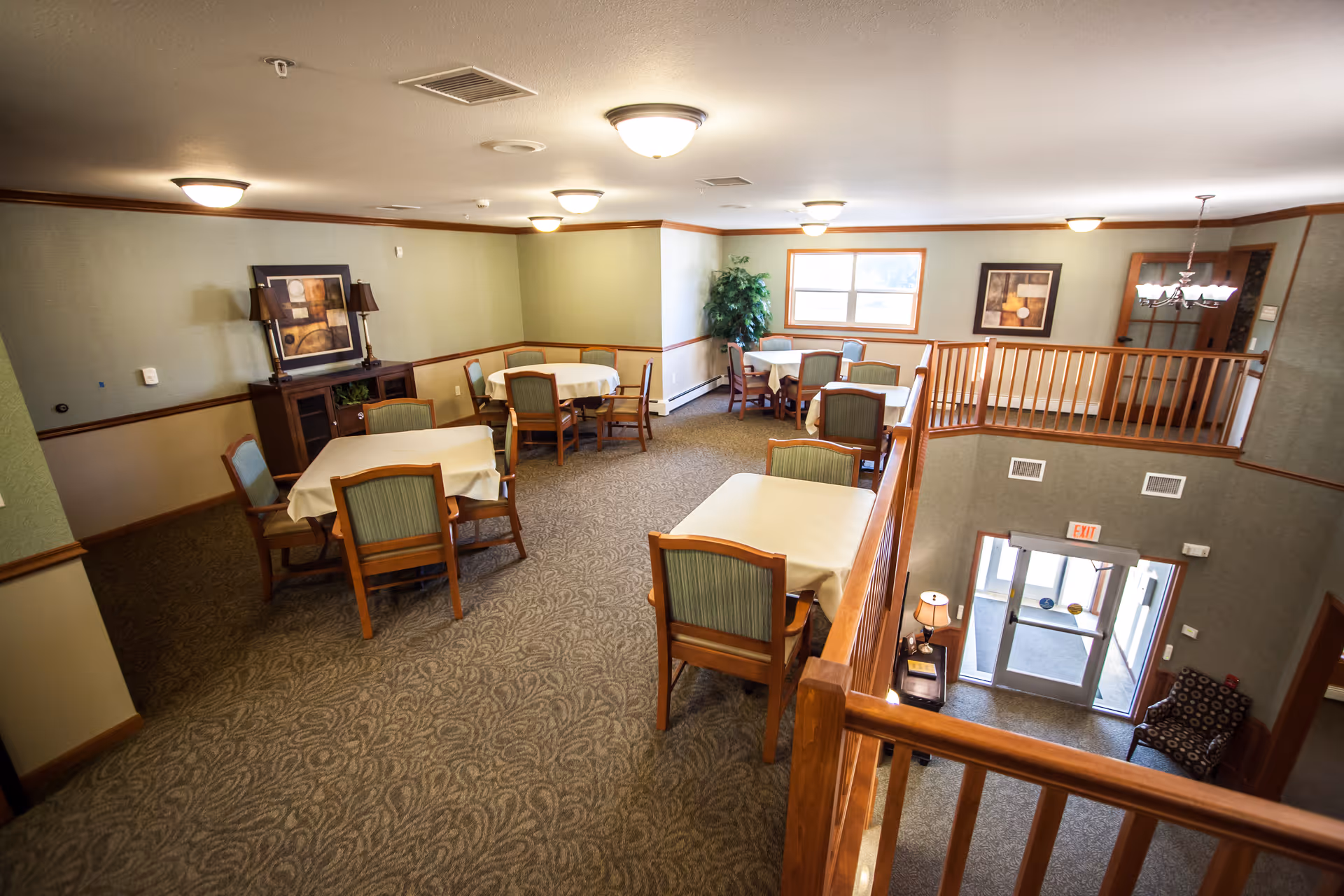 Interior view of a senior living facility dining area with multiple tables covered with white tablecloths and wooden chairs with green upholstery. The room has beige walls with wooden trim, carpeted floor, framed artwork on the walls, and ceiling lights. There is a window letting in natural light and a staircase leading down to an entrance door.