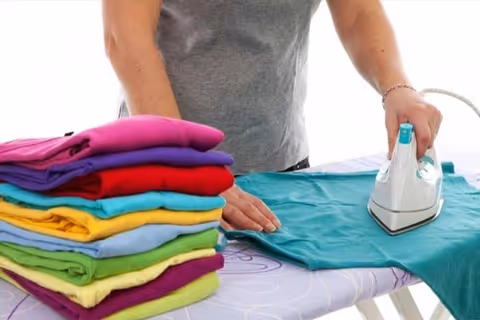 Person irons turquoise fabric on an ironing board beside a stack of colorful clothes