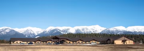 A row of low, single-story buildings sits in a flat field with snow-capped mountains behind.