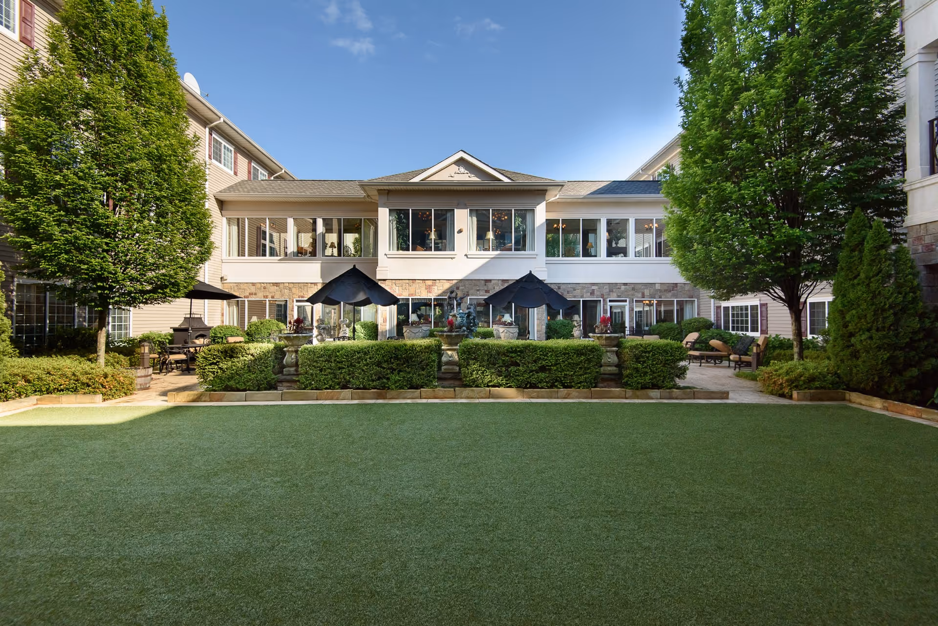 Courtyard lawn and patio with umbrellas and seating in front of a two-story senior living building.