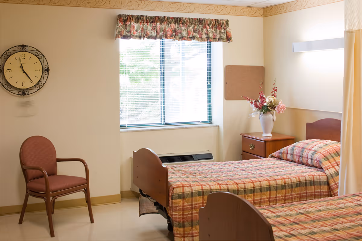 A bright senior living bedroom with two twin beds with plaid bedspreads, a nightstand with a vase of flowers, a chair and a window.