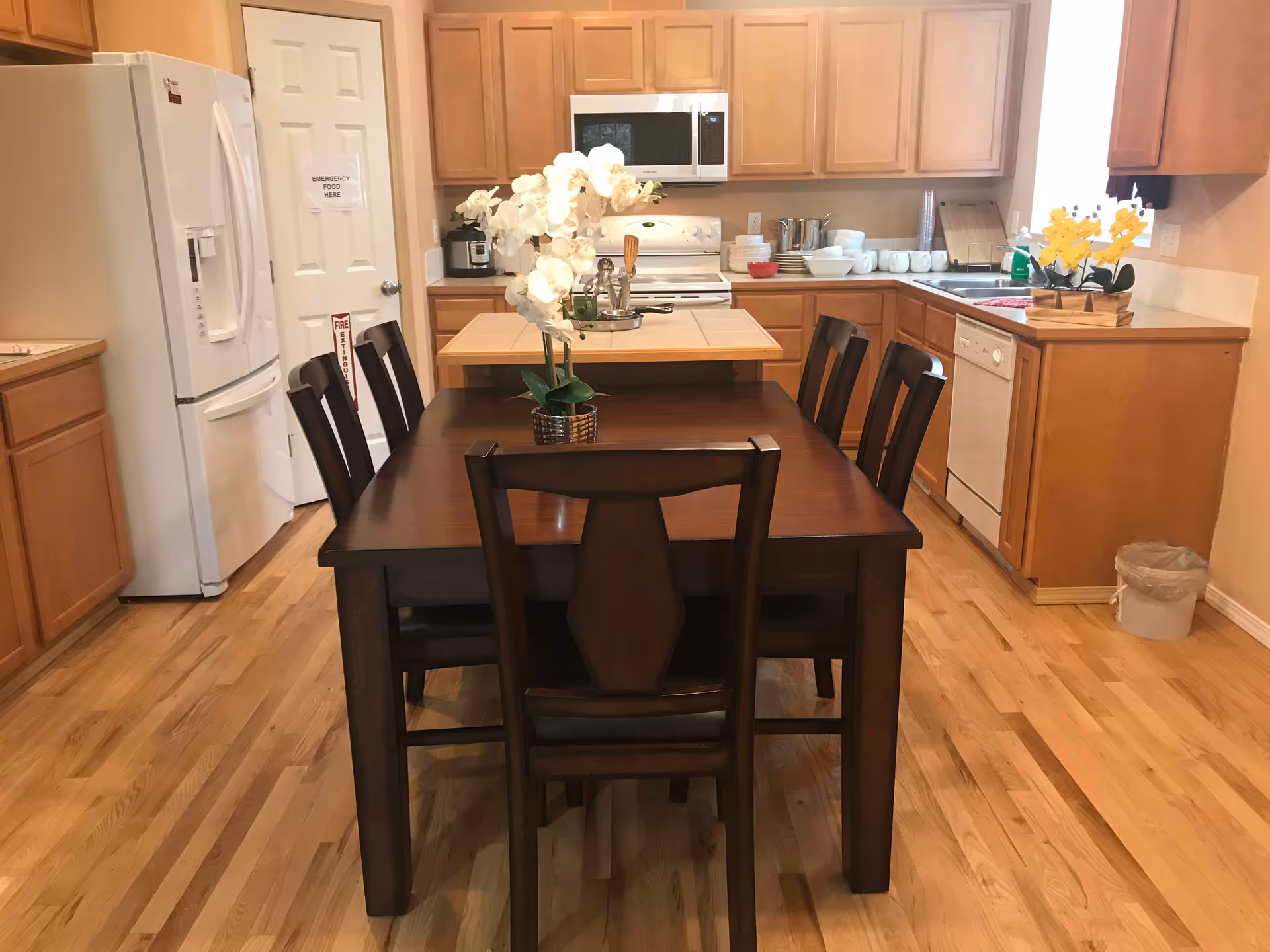 Open kitchen with wooden cabinets and appliances and a dark wood dining table with six chairs in the center.
