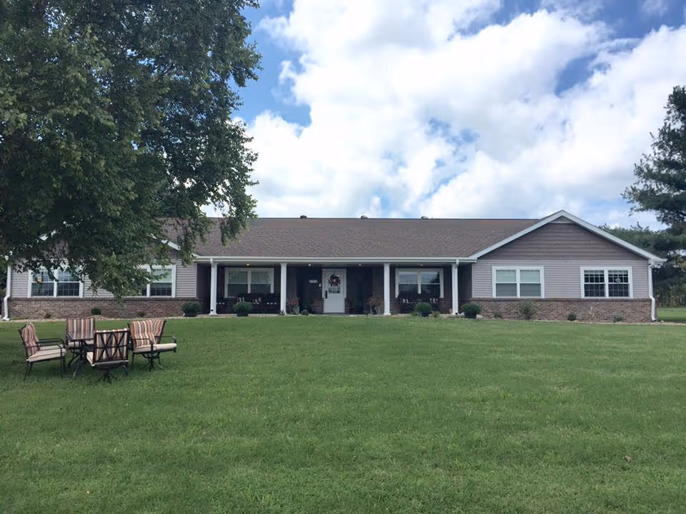 Single-story building with a covered front porch, wide green lawn and outdoor chairs under a partly cloudy sky.