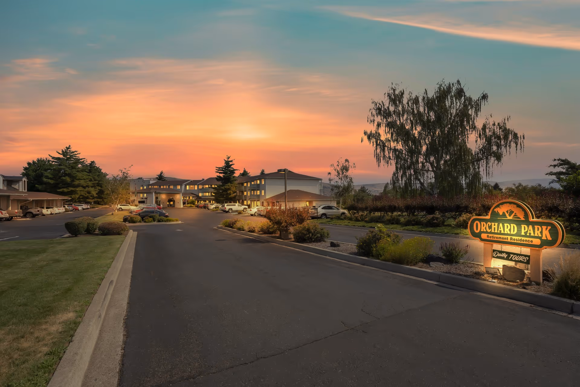 View of a retirement residence building at sunset with a parking lot in front and a sign that reads 'Orchard Park Retirement Residence Daily Tours'. The sky is colorful with shades of orange and pink, and there are trees and landscaped bushes around the area.