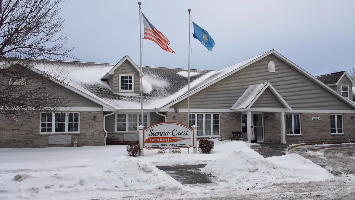 Exterior view of Sienna Crest Assisted Living, Inc. building in winter with snow on the ground and roof. Two flagpoles with the American flag and another flag stand in front of the building near a sign displaying the facility's name and phone number.