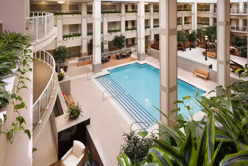 Indoor atrium featuring a rectangular swimming pool surrounded by seating areas, plants, and multiple balcony levels.