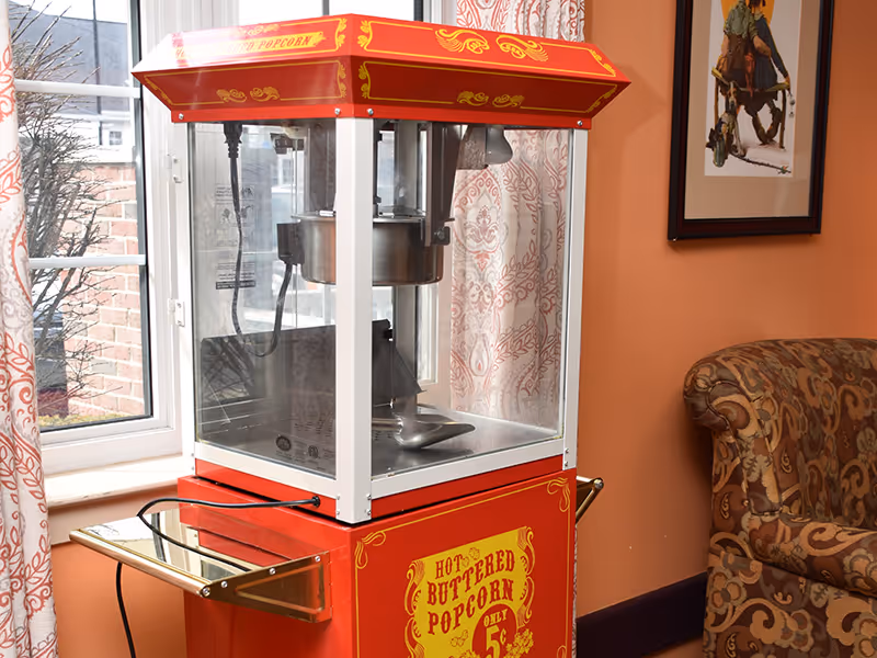 Red vintage popcorn machine sits indoors by a window next to a patterned armchair and framed artwork.