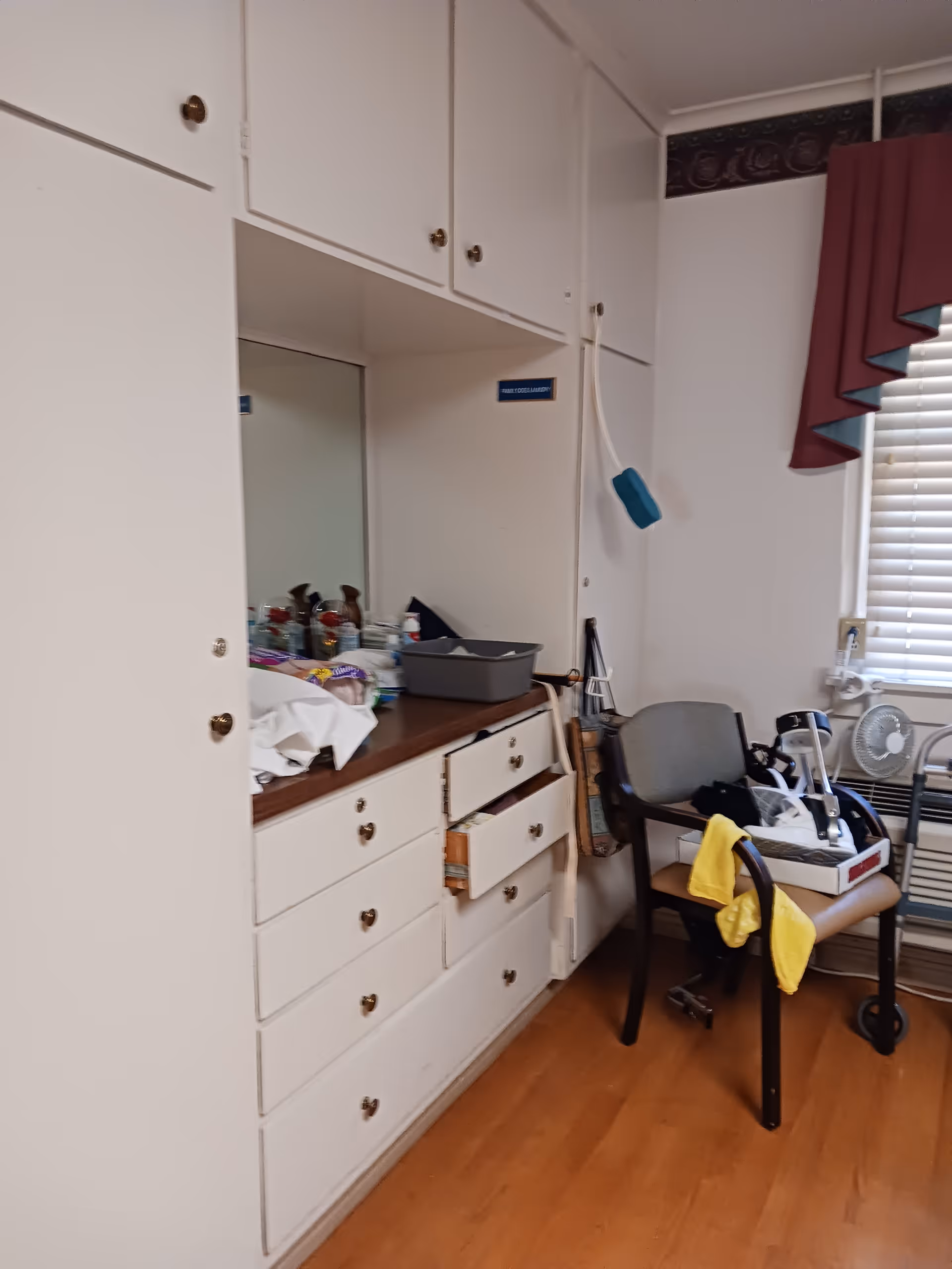 A cluttered senior care room with white cabinets, a countertop, and a chair by a window.