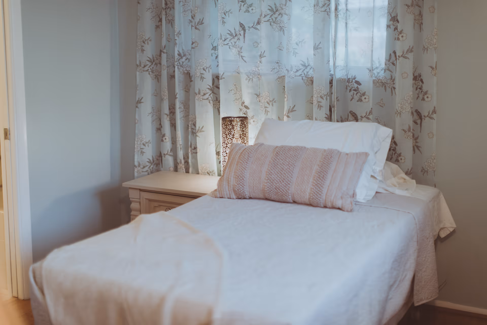 A neatly made bedroom with a bed, floral curtains, and a nightstand lamp.