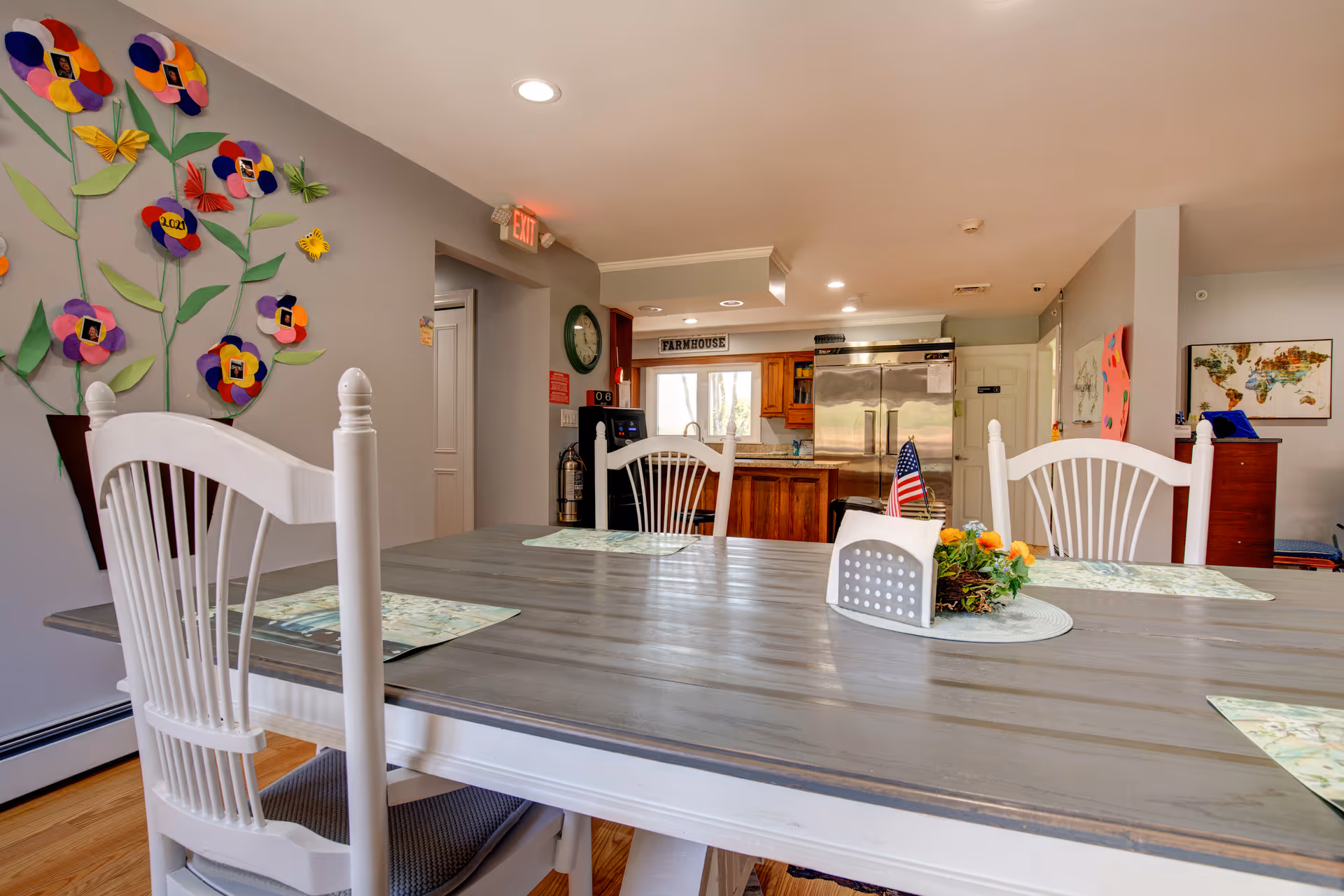 Bright dining area with a long table, white chairs, and a kitchen visible in the background.