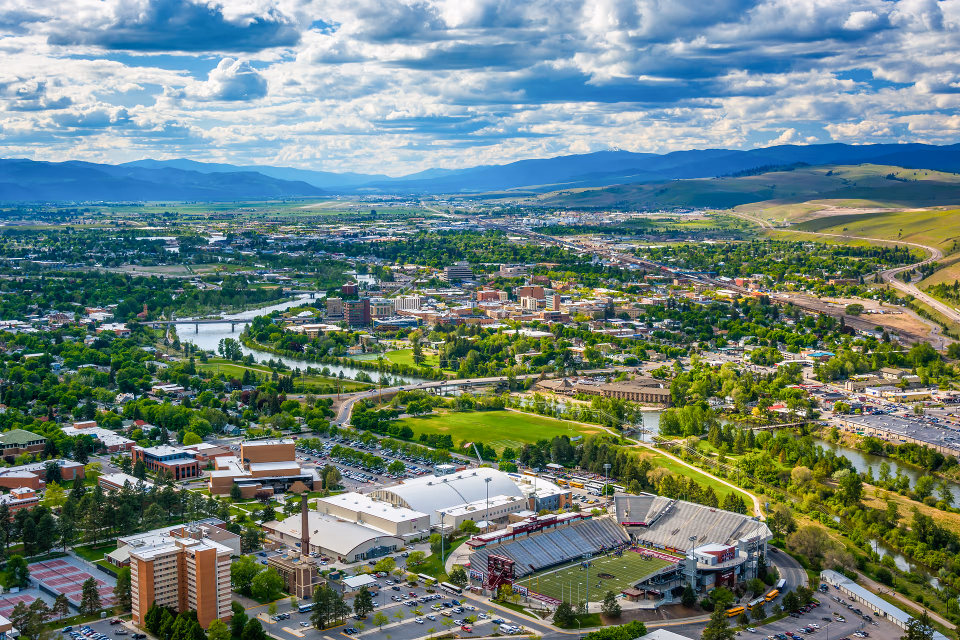 Aerial view of a suburban cityscape with a river, hills, and a stadium.