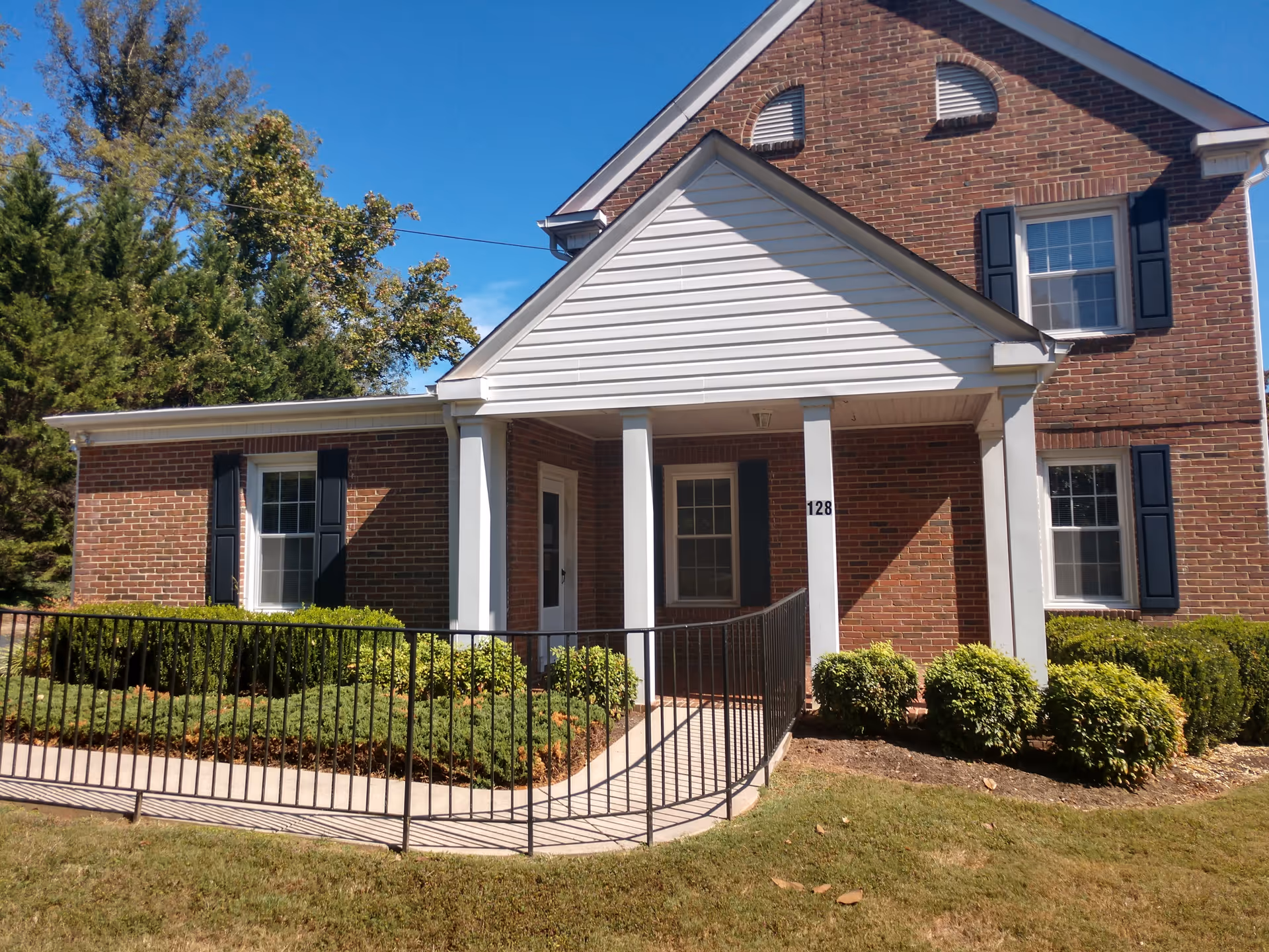 Brick building with white columns and a ramped entrance, surrounded by shrubs.