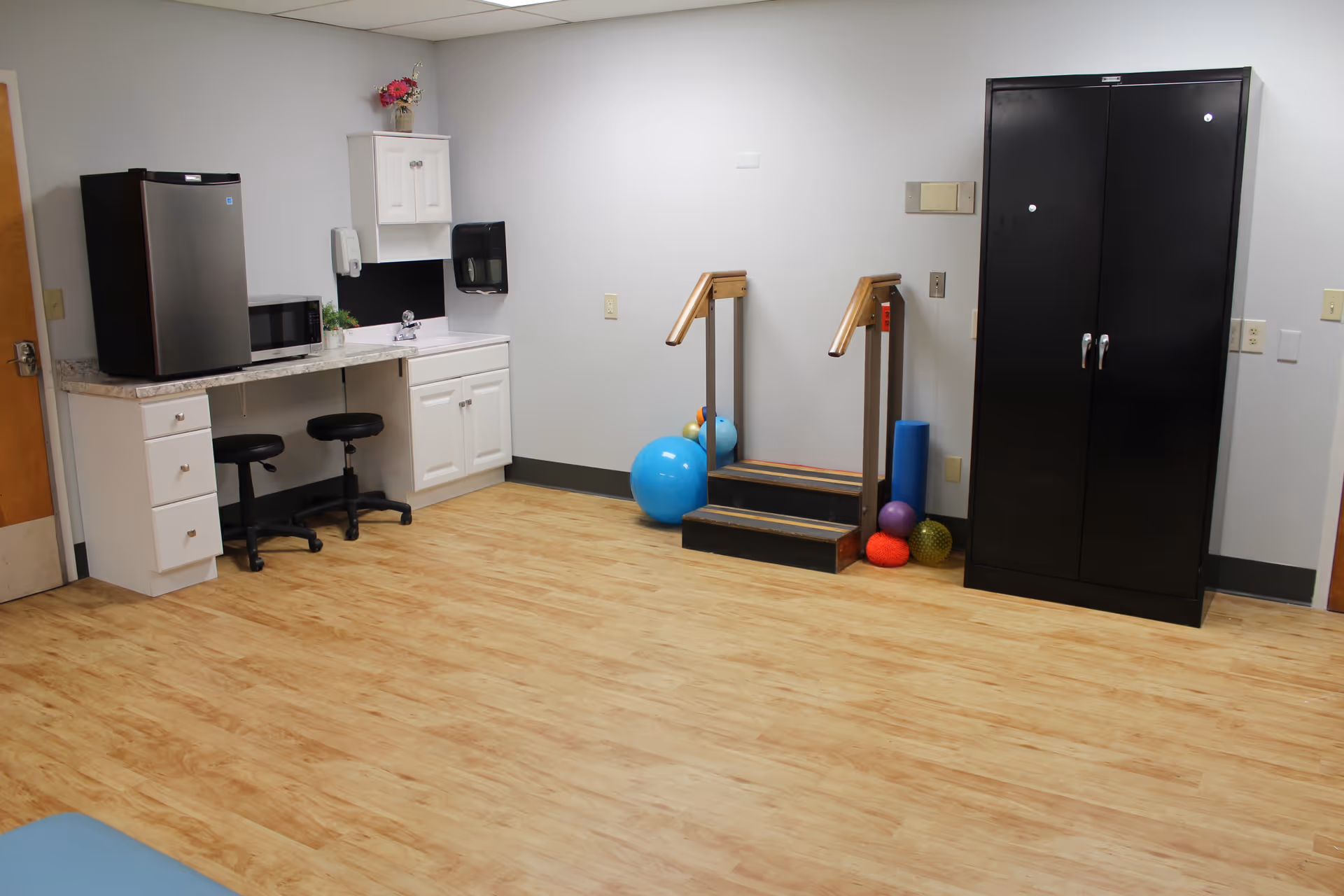A room with light wood flooring and white walls featuring a small kitchenette area with a mini fridge, microwave, two black stools, and white cabinets. There is a small set of wooden stairs with handrails and various exercise balls and equipment nearby. A tall black cabinet stands against the wall on the right side.