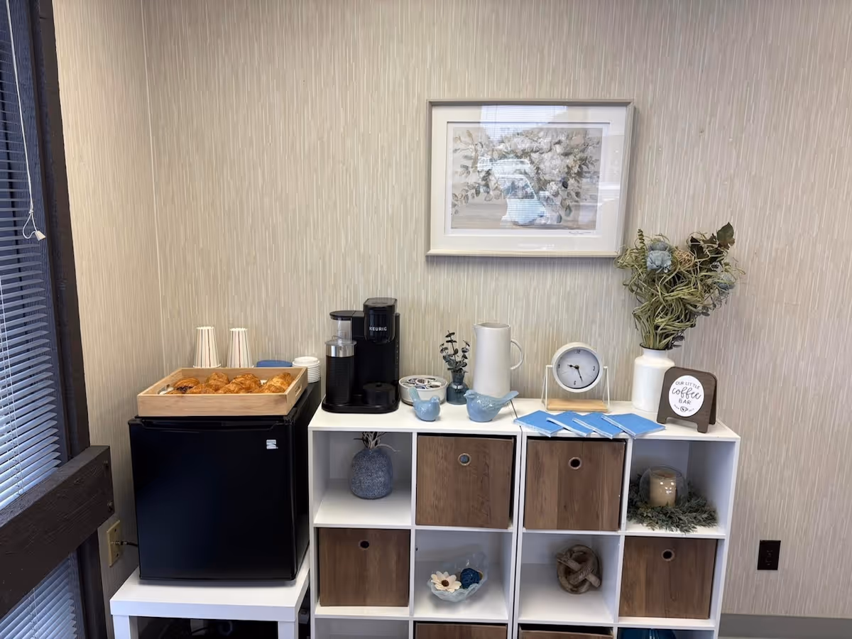 Coffee station with a Keurig, pastries, mugs, framed art, and cubby shelves in a small lounge.