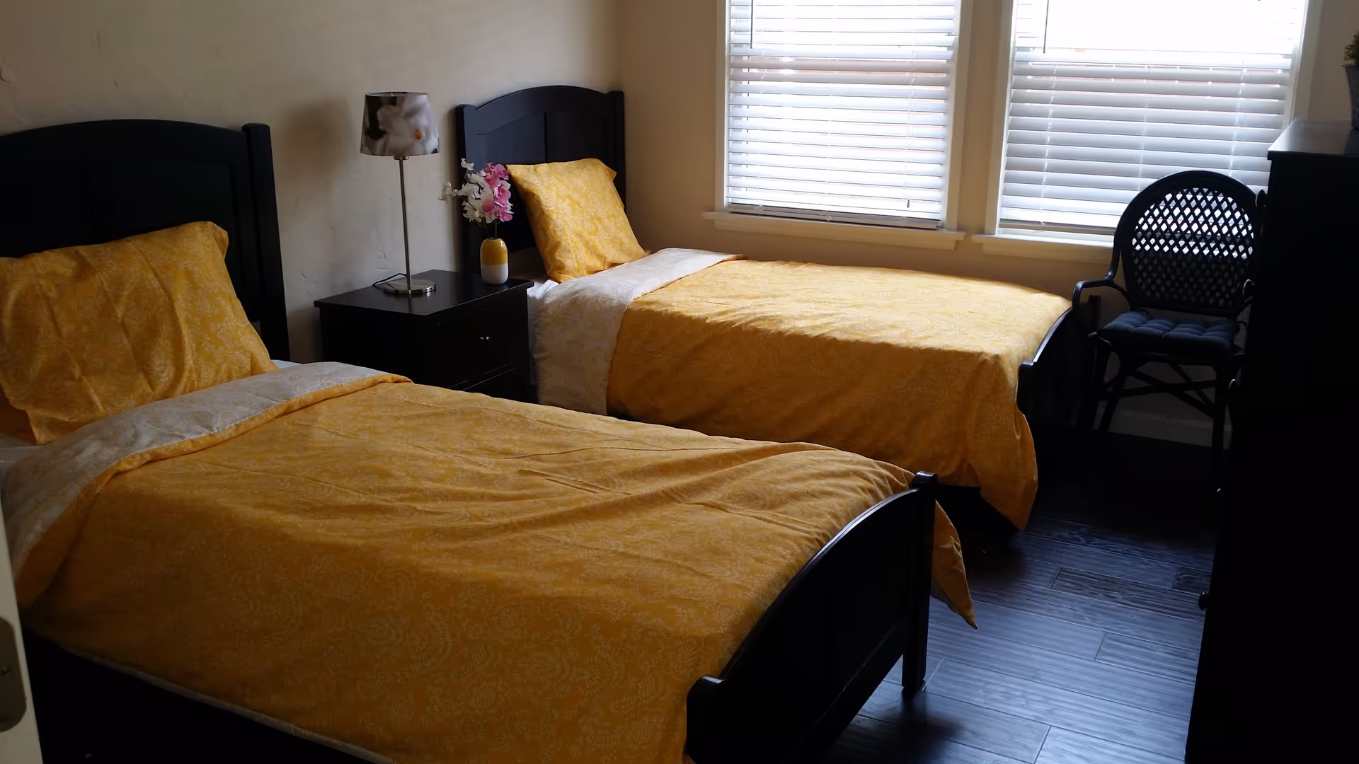 A bedroom with two single beds featuring yellow patterned bedspreads and pillows. Between the beds is a dark wooden nightstand with a floral lamp and a small vase with flowers. The room has two windows with white blinds, a dark wooden dresser, and a black chair with a cushion.