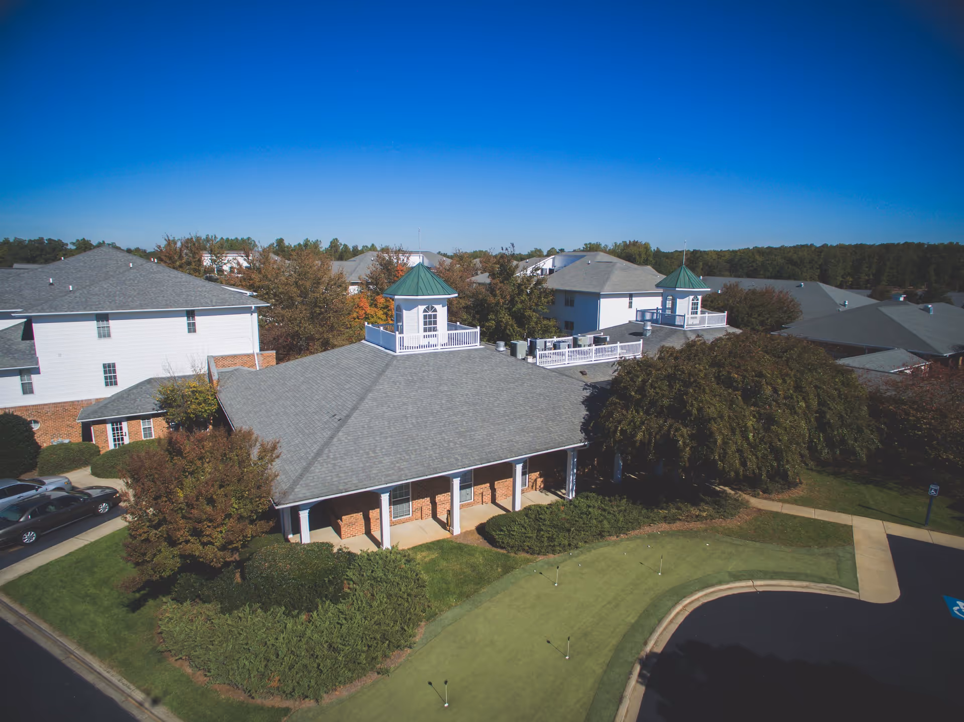 Aerial view of a senior living facility named Summit Hills showing multiple connected buildings with gray roofs and white walls, surrounded by trees and greenery under a clear blue sky. There is a small putting green and a parking area with cars visible in the foreground.