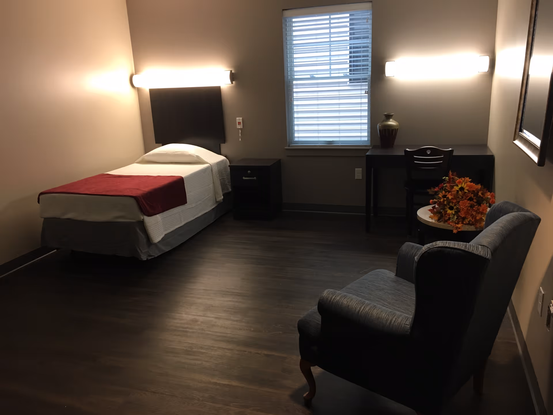 A simple bedroom with a single bed covered with white bedding and a red throw blanket. There is a nightstand next to the bed, a window with blinds, a desk with a chair and a decorative vase, and a small round table with a flower arrangement. A cushioned armchair is positioned near the table. The room has dark wood flooring and neutral-colored walls with two horizontal wall lights above the bed and desk.