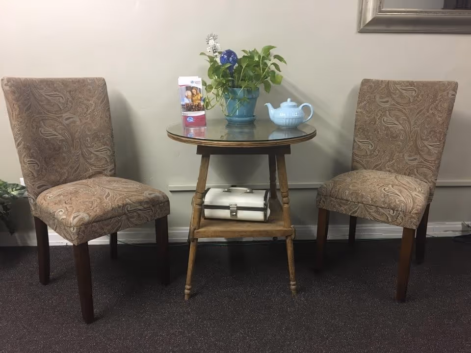 Two upholstered chairs flank a round glass table with a plant and teapot.
