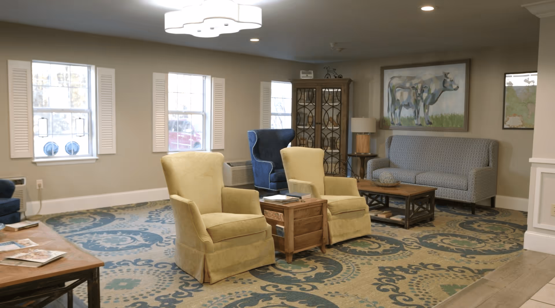 A cozy living room area with two yellow armchairs, a blue armchair, and a patterned blue sofa. The room features a wooden coffee table, a side table with a lamp, a patterned carpet, and three windows with white shutters. There is a large painting of a cow on the wall and a glass-front cabinet in the corner.