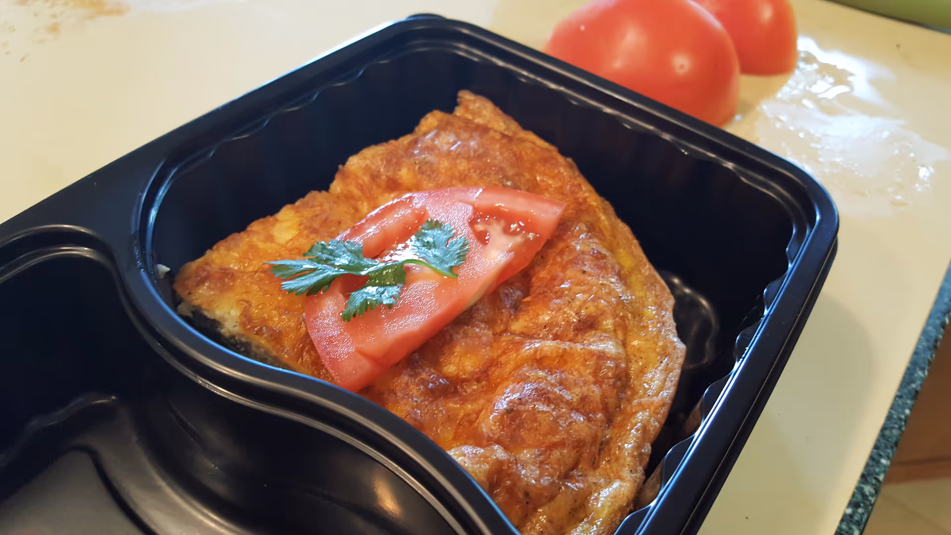 A golden baked dish in a black takeout tray with tomato garnish on a light countertop.