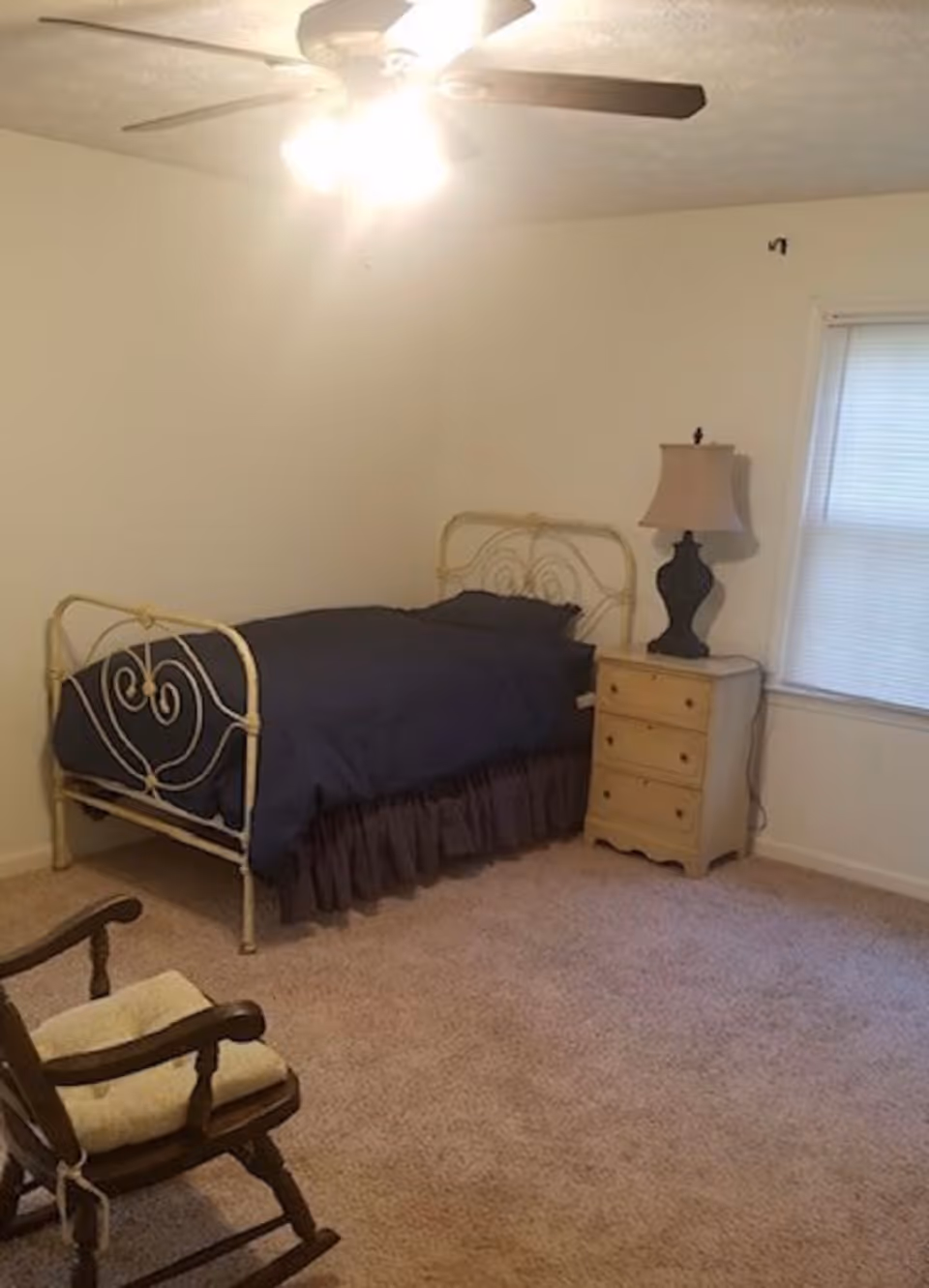 Small carpeted bedroom with a metal-frame twin bed covered in dark bedding, a nightstand with a lamp, a rocking chair, and a ceiling fan.