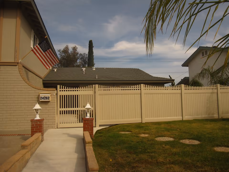 Front exterior of a house showing a walkway, brick pillars with lamps, a gated beige fence, an American flag, and house number 24262.