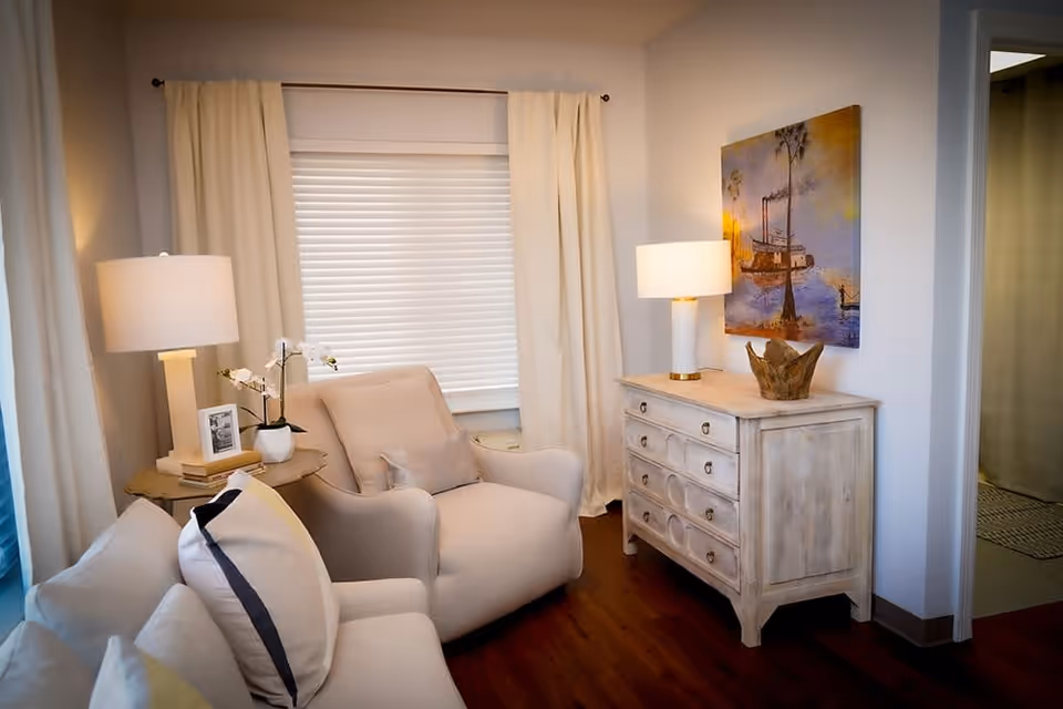 A cozy living room area with a beige armchair and a white sofa adorned with pillows. There are two table lamps on either side, one on a small round side table with a framed photo and a small plant, and the other on a wooden chest of drawers. A colorful painting hangs on the wall above the chest. The room has white curtains and a window with blinds, and the floor is wooden.