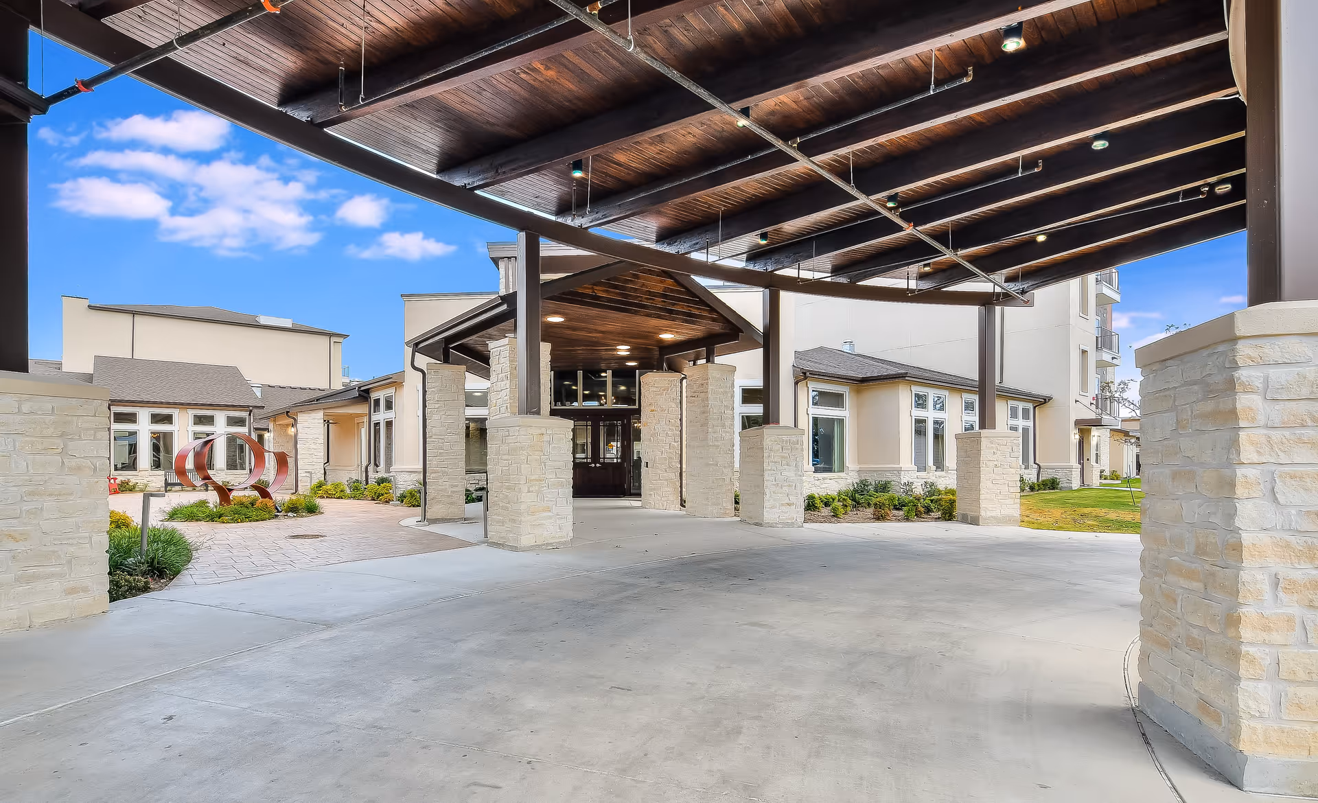 Covered entrance area of a senior living facility with stone pillars and a wooden ceiling, leading to the main building entrance. The exterior features beige walls, multiple windows, and landscaped greenery with a decorative sculpture on the left side.