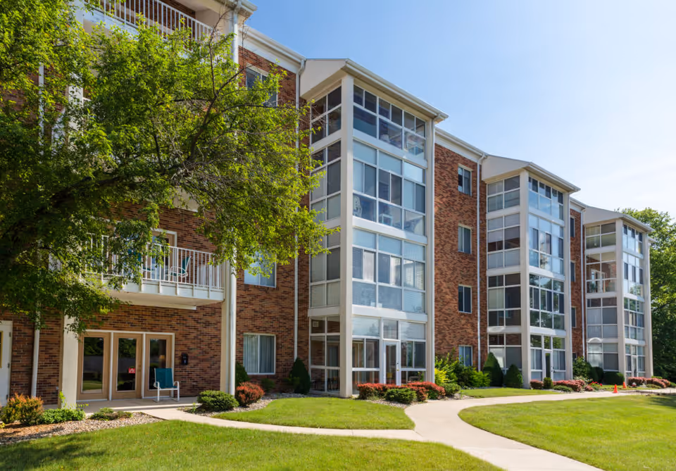 Exterior of a multi-story brick senior living building with glass-enclosed balconies, a curved walkway and lawn.