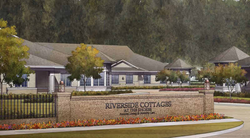 Brick entrance sign reading 'Riverside Cottages at the Shores' with landscaped flowerbeds and a single-story cottage-style building behind it.