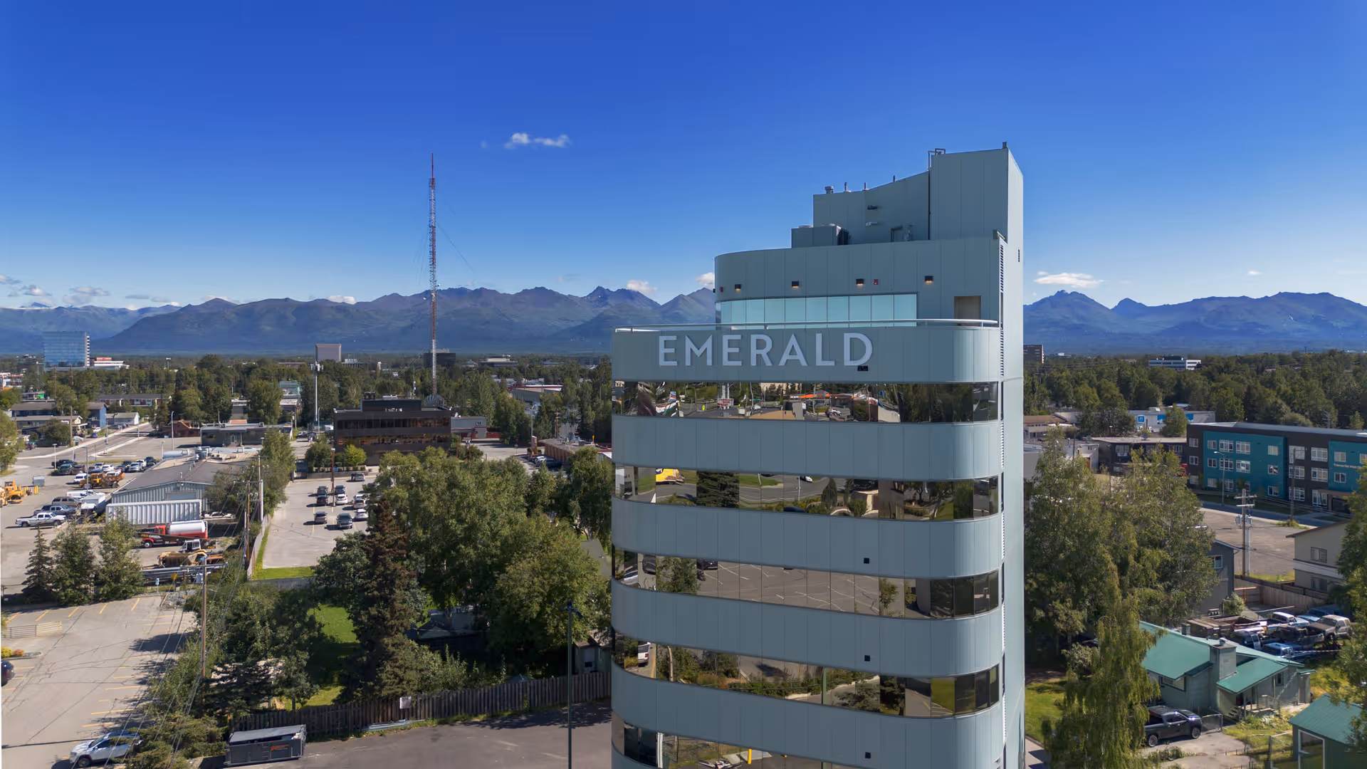 A tall modern building with reflective windows and the word 'EMERALD' displayed near the top, surrounded by trees, parking lots, and other buildings with mountains visible in the background under a clear blue sky.