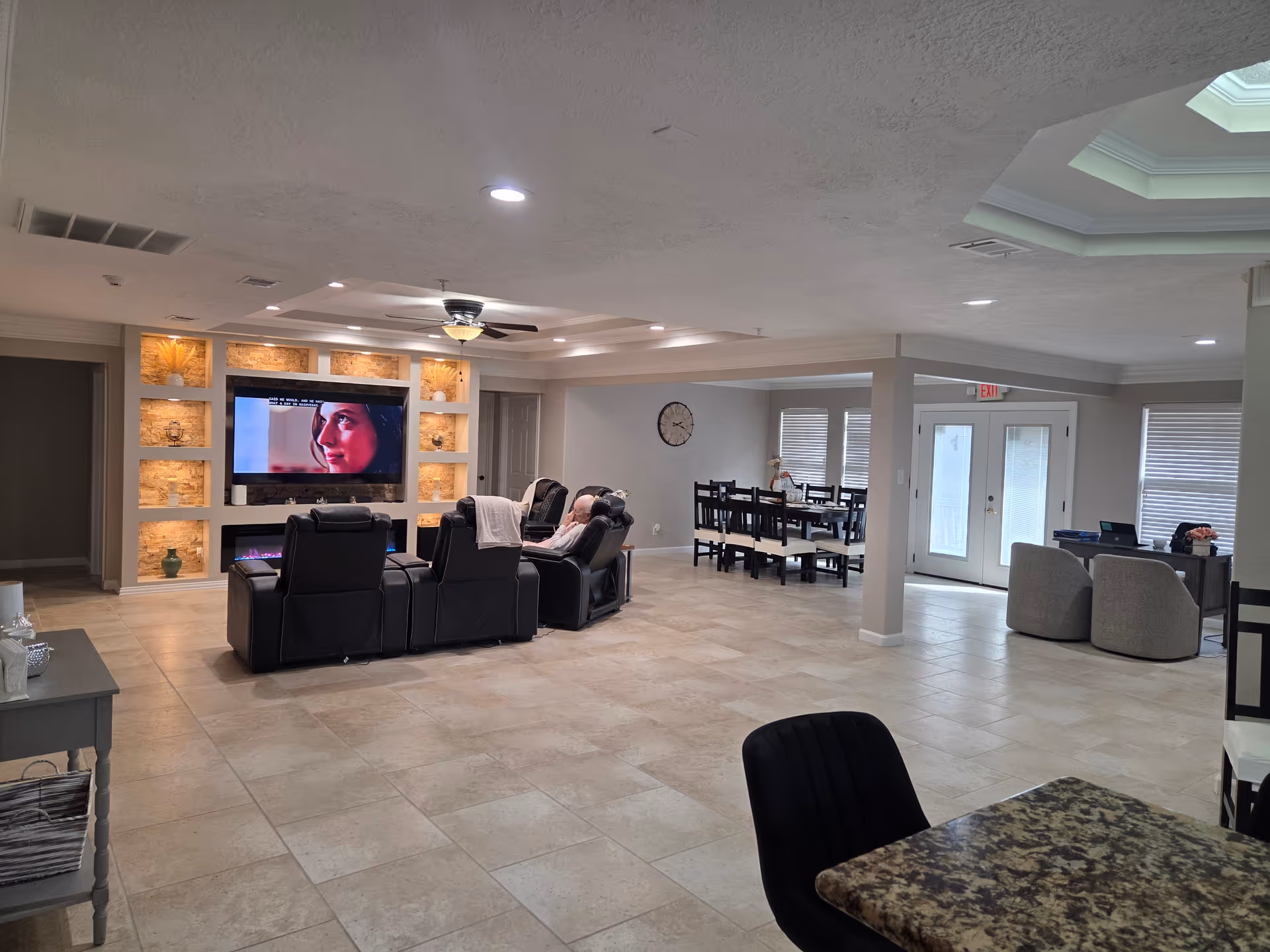 Spacious senior living lounge with wall-mounted TV, recliners, dining area, and entry doors.