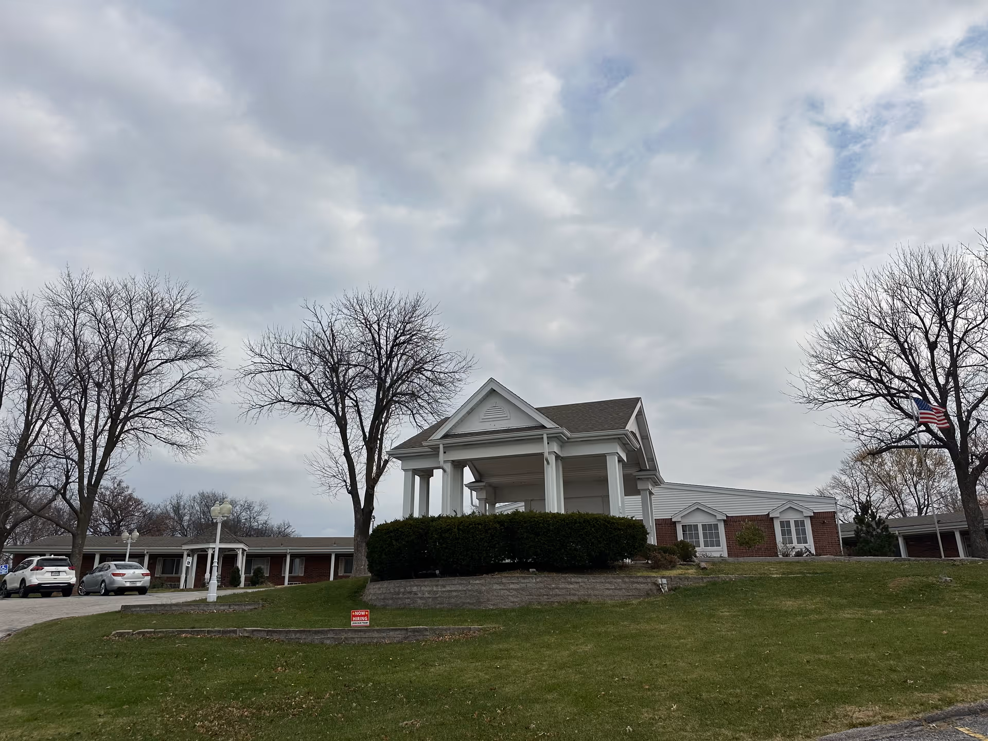 Front exterior of a single-story red-brick senior living facility with a white columned entrance, lawn, bare trees, parked cars, and an overcast sky.