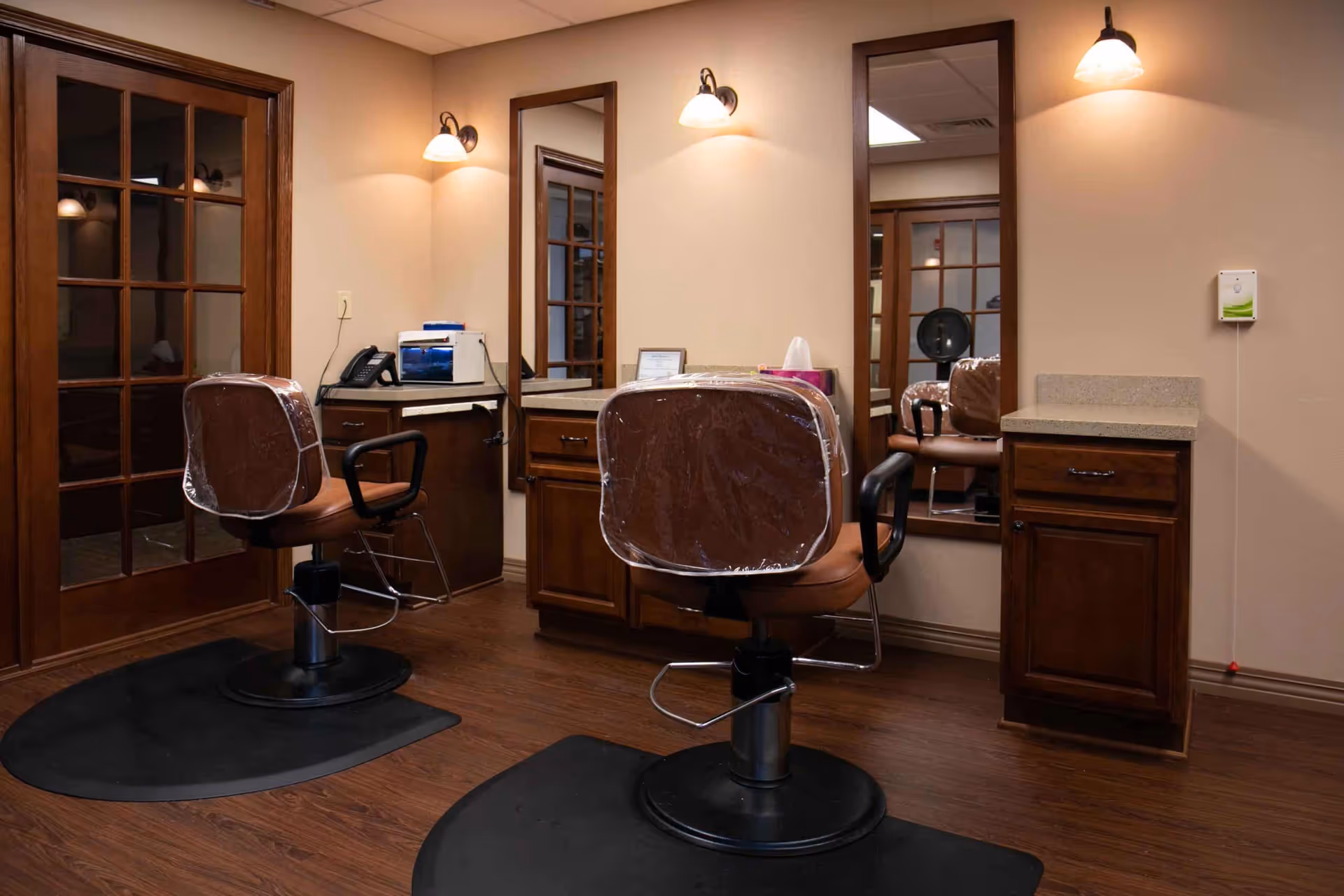 Interior view of a senior living facility's salon area with two brown salon chairs covered in plastic, positioned in front of large mirrors mounted on the wall. The room has wooden flooring, wooden cabinetry with countertops, wall-mounted lights above each mirror, and a set of glass-paneled double doors.