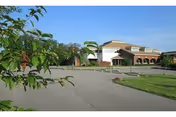 Exterior view of a senior living facility with entrance, greenery, and parking.