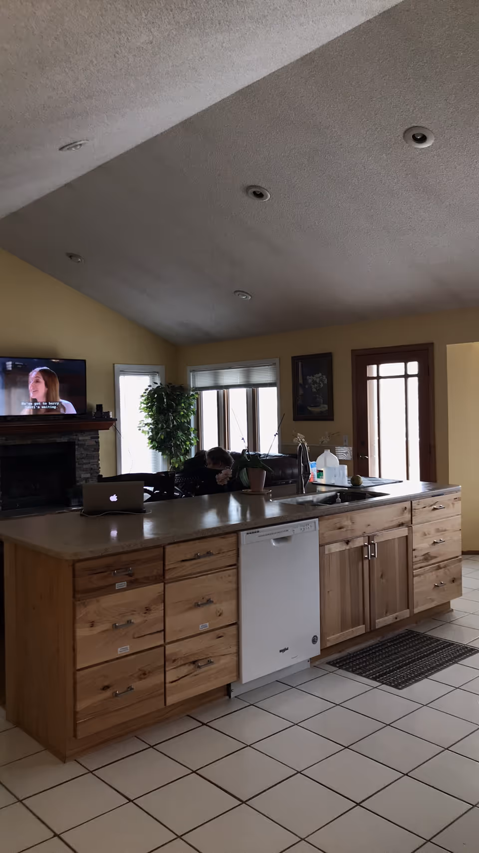 Open-concept kitchen with wooden cabinets, central island, dishwasher, and TV near a fireplace.