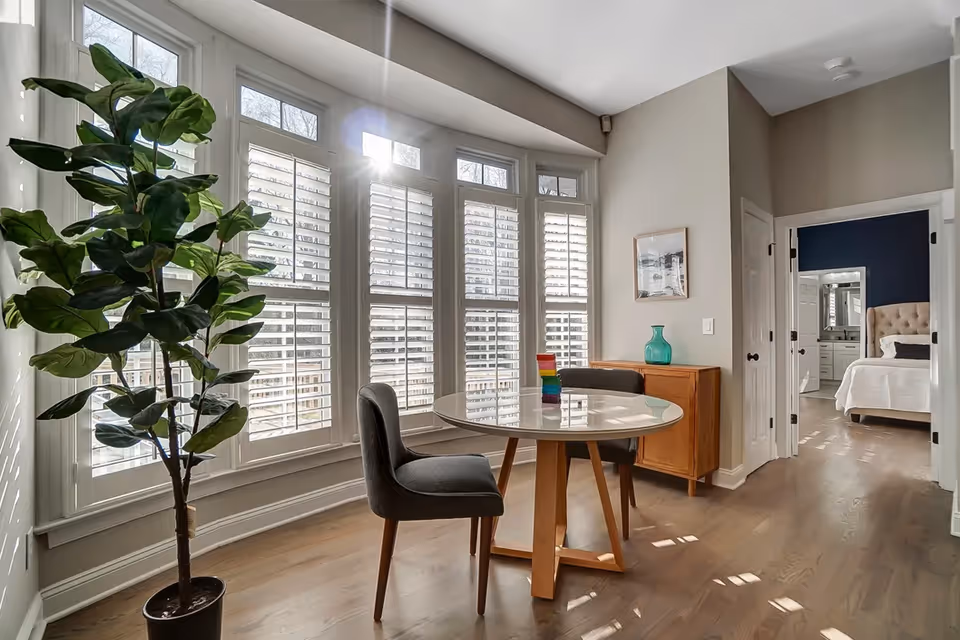 Bright corner interior with a round glass table, two chairs, tall plant, and large bay windows.