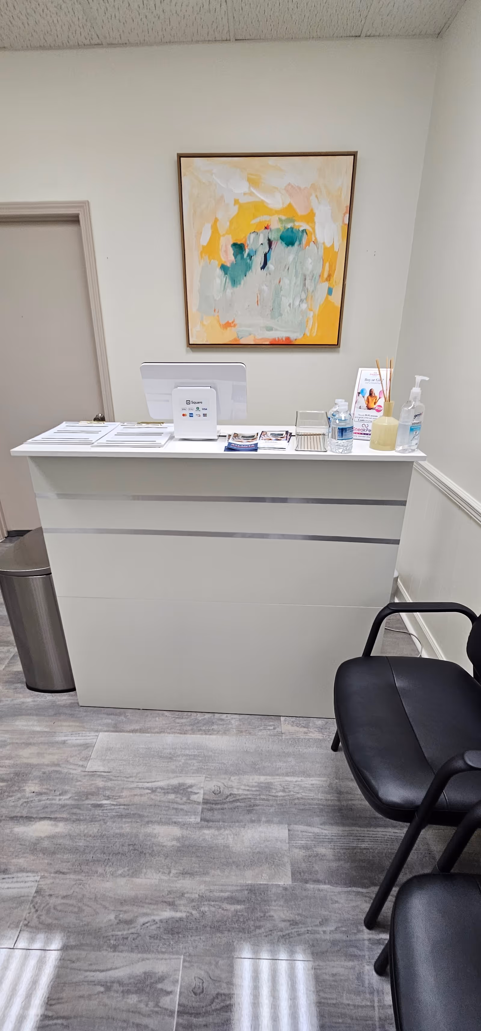 Modern reception desk with abstract painting, sanitizer, and black waiting chairs in a clean facility lobby.