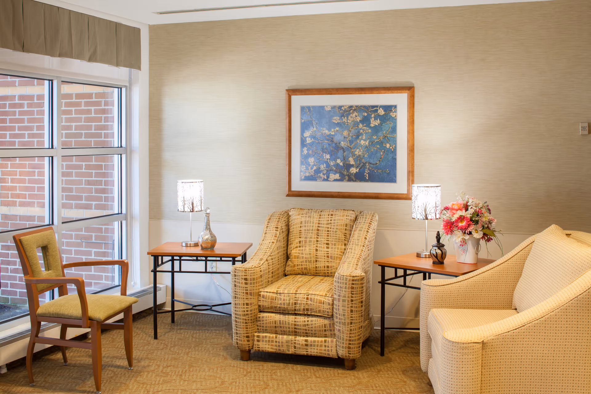 A cozy sitting area in a senior living facility with a large window on the left showing a brick wall outside. Inside, there is a wooden chair with green upholstery, a patterned armchair, and a cream-colored sofa. Two wooden side tables hold decorative lamps and a vase with pink and white flowers. A framed artwork featuring a floral design hangs on the beige wall behind the armchair.