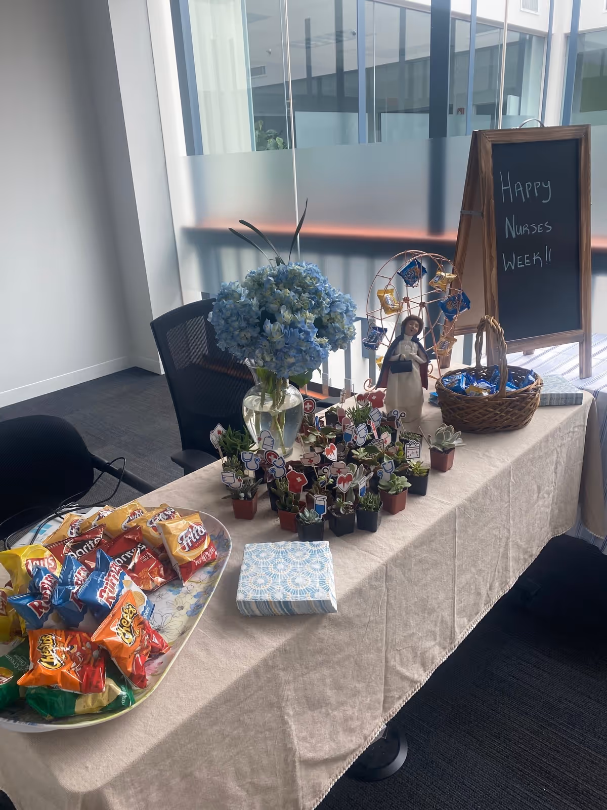 Indoor event table with snacks, potted plants, a chalkboard sign honoring Nurses Week near glass wall.