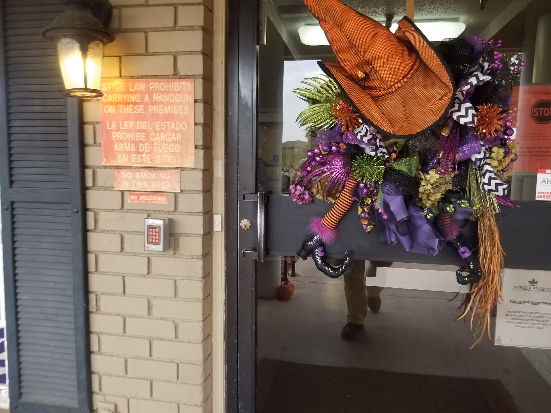 Colorful wreath decorates a glass door at a building entrance with brick wall and lantern.