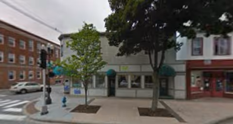 Street view of a storefront row with trees, awnings, and a crosswalk along the sidewalk.