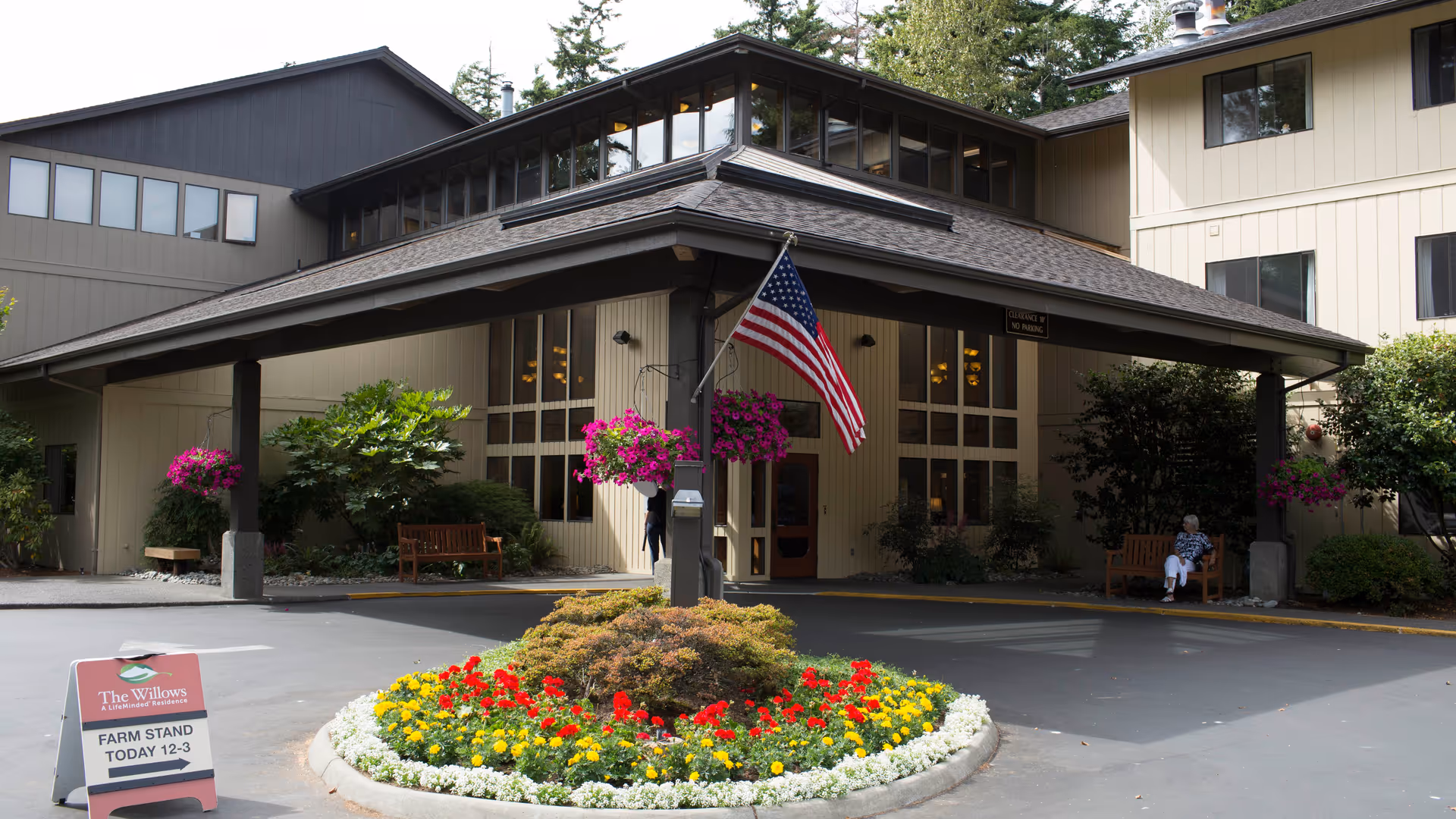 Exterior view of The Willows Bellingham entrance with flag, benches, and colorful flower beds.