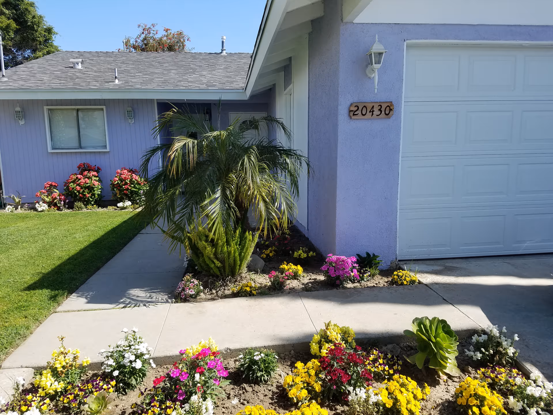 Front exterior view of a single-story house with a light purple exterior wall, a white garage door, and a wooden house number plaque displaying 20430. The front yard features a well-maintained lawn, a concrete walkway, and colorful flower beds with various blooming flowers and green plants.