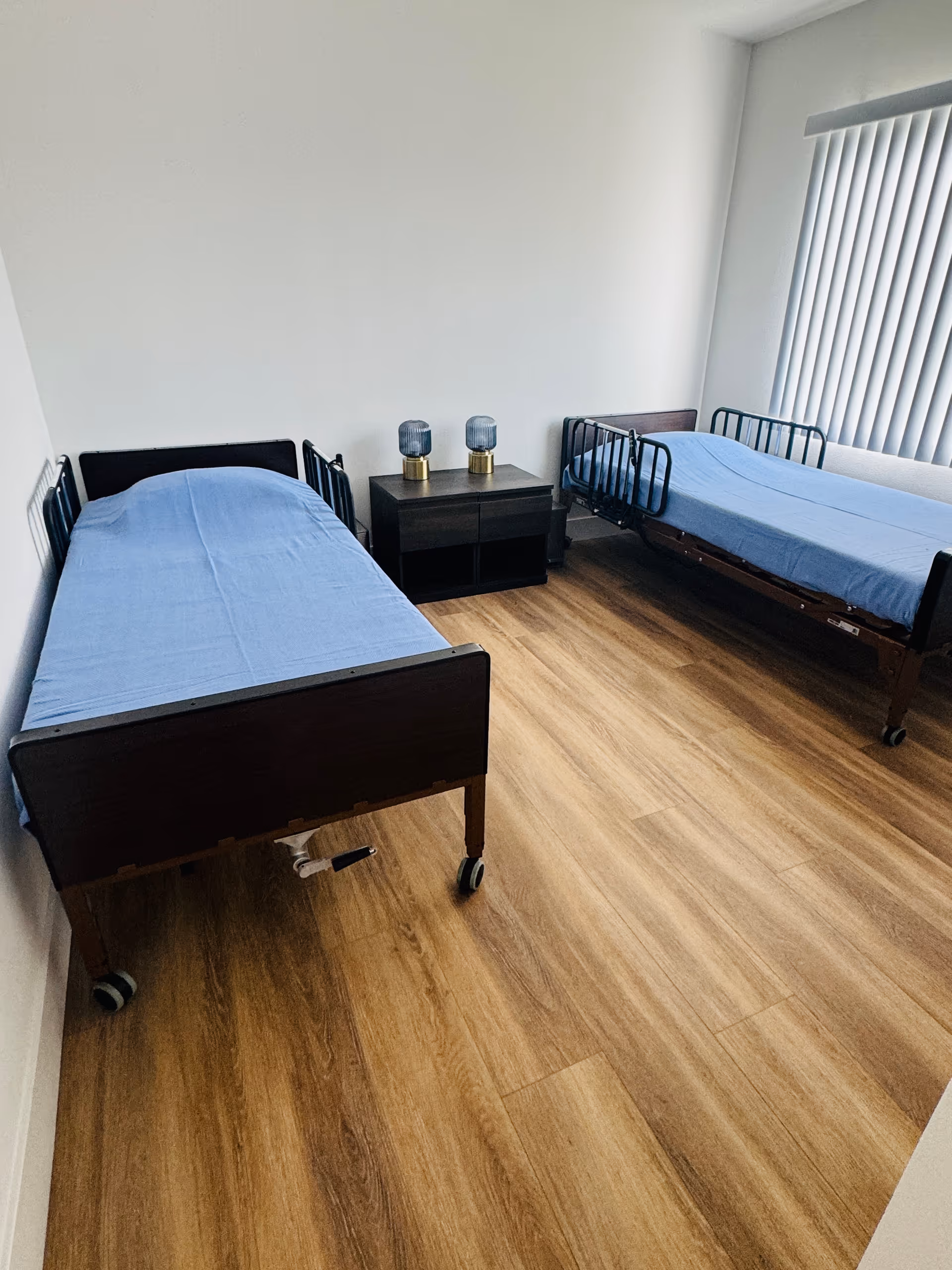 Two twin beds with blue sheets, a shared nightstand, and window blinds in a bedroom.