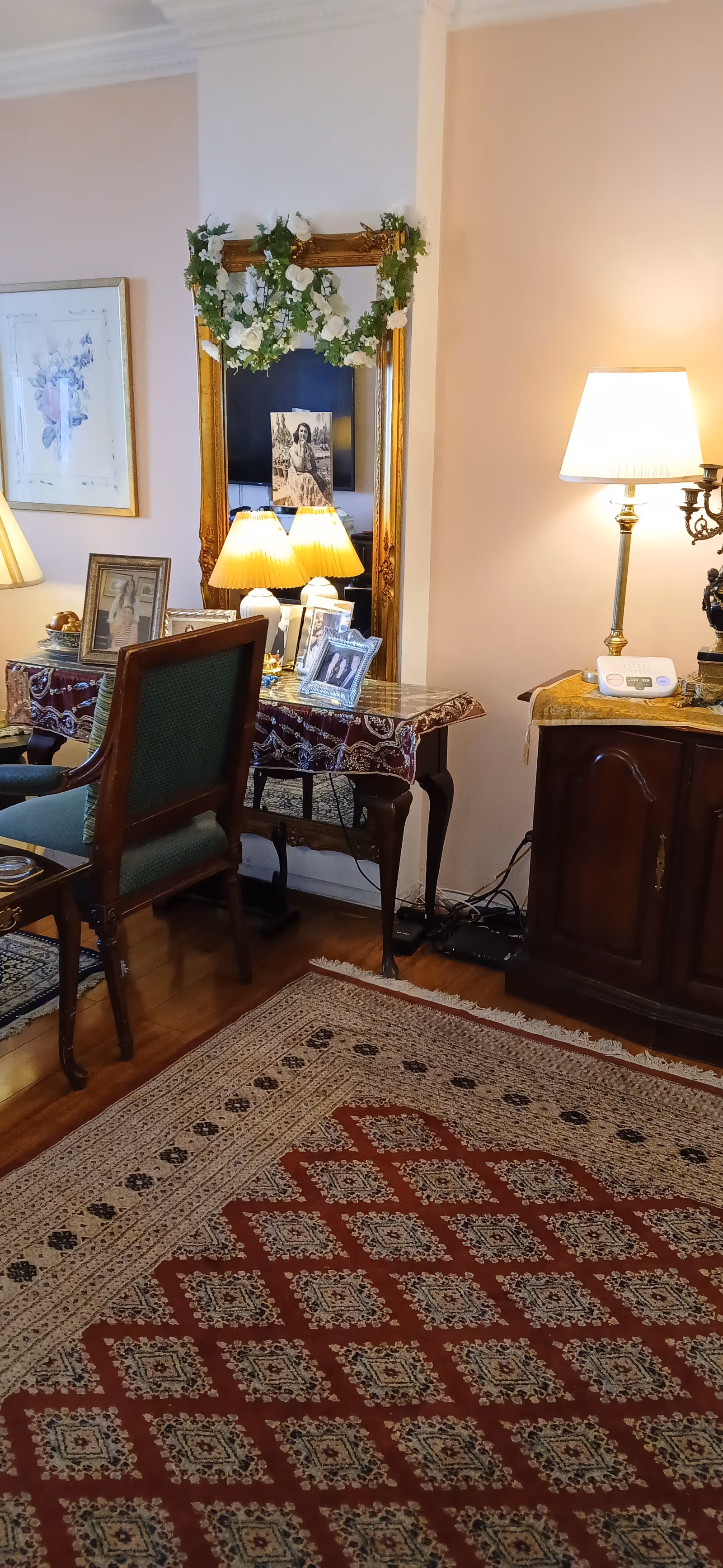 Cozy living room with chairs, lamps, framed photos, and a floral garland around a mirror.