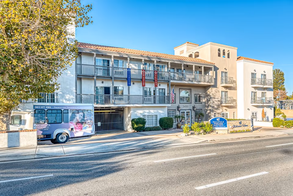 Front exterior of a three-story Mediterranean-style senior living building with balconies, an entrance sign, and a parked shuttle bus.