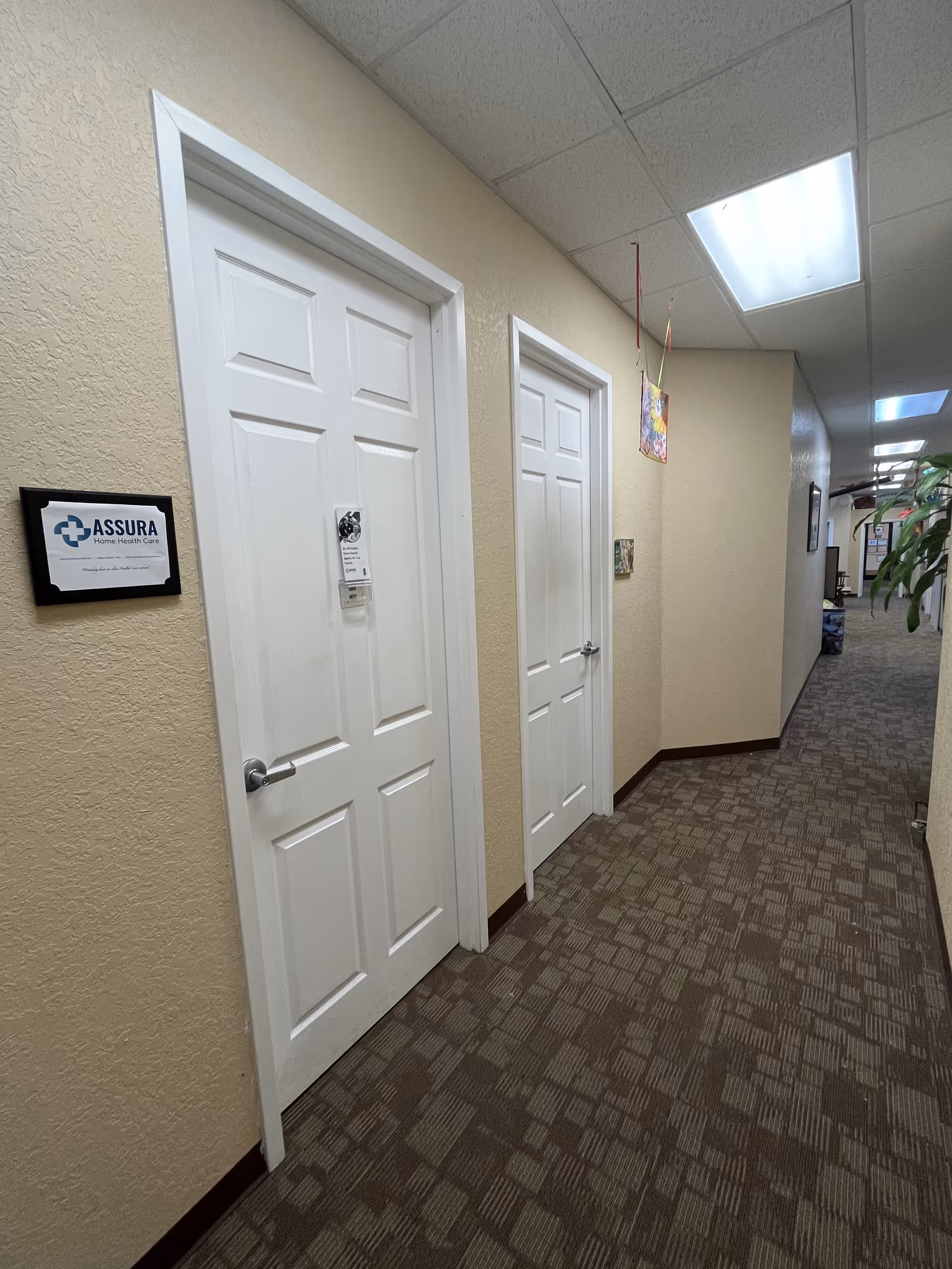 Narrow beige hallway with white doors, ceiling lights, and Assura wall plaque.