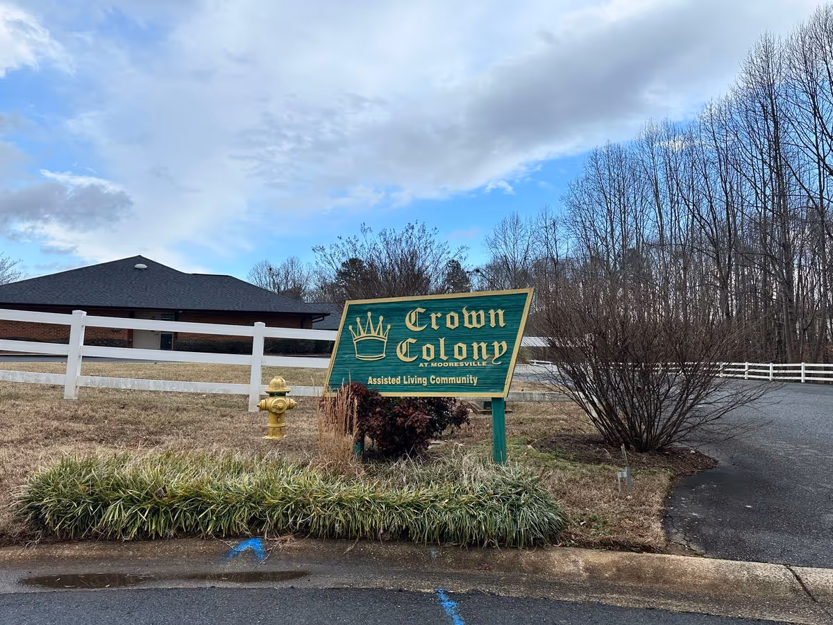 Green "Crown Colony" assisted living community sign in front of a low brick building, white fence, and trees under a cloudy sky.